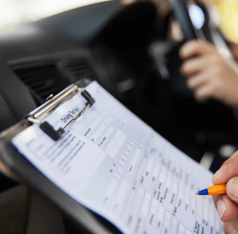 Driving test form on a clipboard in a car as an examiner marks scores while the driver holds the steering wheel.