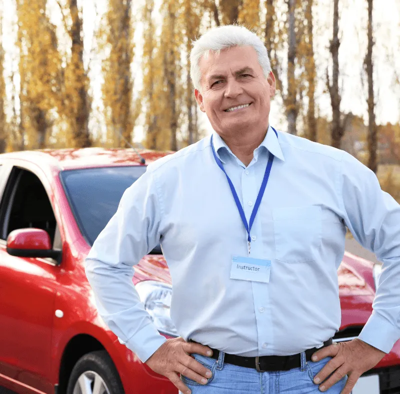 Smiling older male driving instructor wearing a blue lanyard badge standing in front of a red car with autumn trees in background.