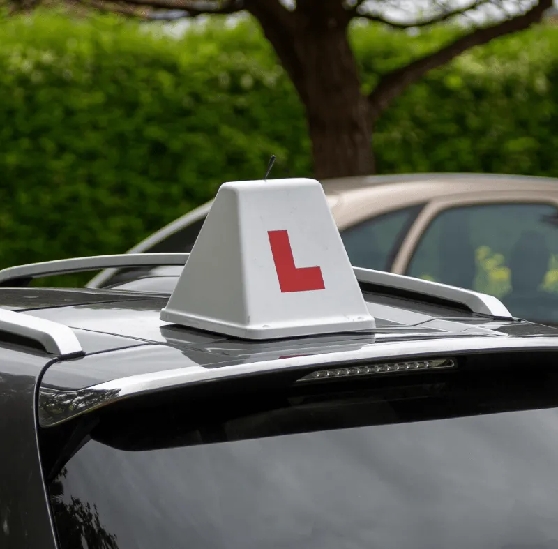 Driving school car roof with a white learner sign showing a red “L” against a leafy background.