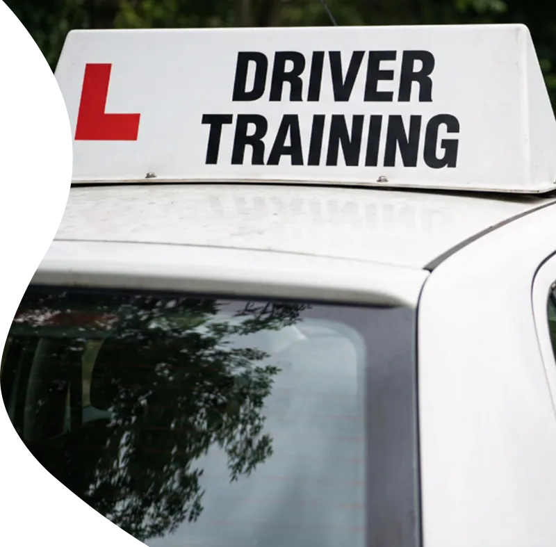 White car with a rooftop sign reading “Driver Training” and a red learner L plate, parked near trees.