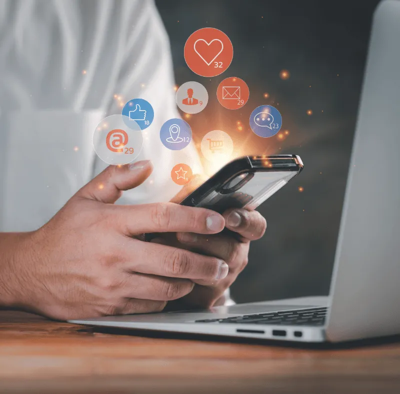 Hands holding a smartphone above a laptop, with glowing social media notification icons floating from the screen.