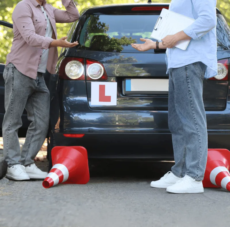 Learner driver and driving instructor standing beside a car with L plates and knocked-over cones after a dual control driving lesson accident.