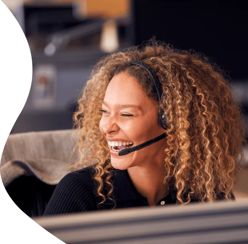 Smiling call center agent with curly hair wearing a headset, laughing while working at her desk.