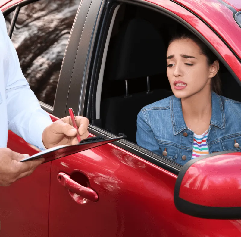 Worried learner driver sitting in a red car while an examiner writes notes on a clipboard during a driving test.