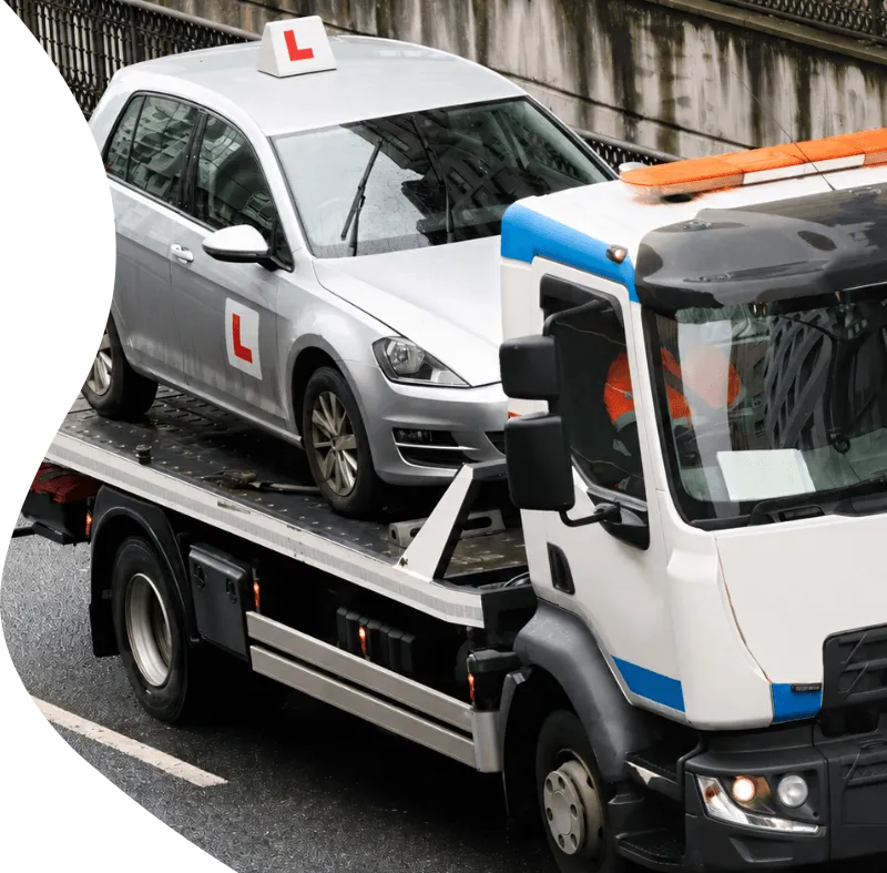 A silver driving school car with L plates being transported on a flatbed breakdown recovery truck.