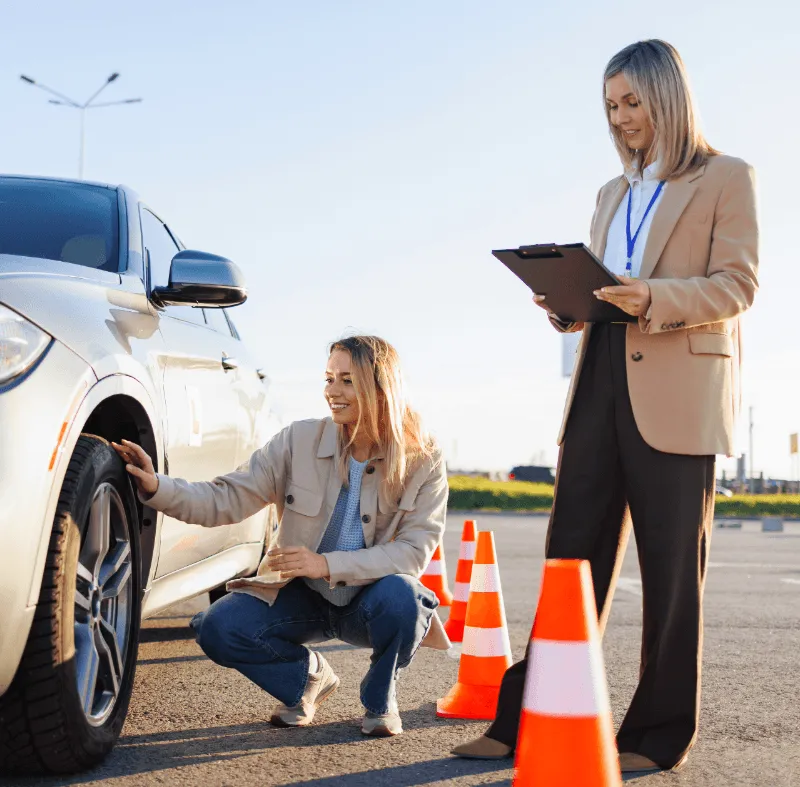 A driving examiner with clipboard watches a young woman inspect a car tire near orange traffic cones during a driving test.