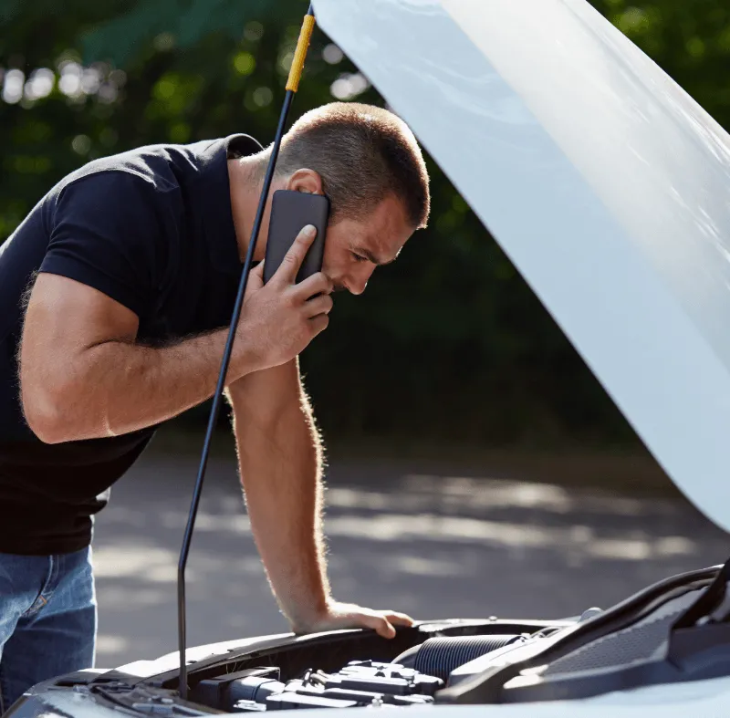 Man in black t-shirt talking on phone while inspecting open car engine hood outdoors, possibly calling for roadside assistance.