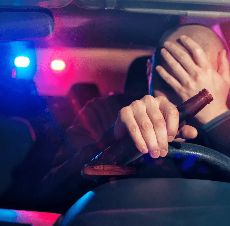 Driver holding a beer bottle at the steering wheel with police lights visible through the rear window, depicting drunk driving.