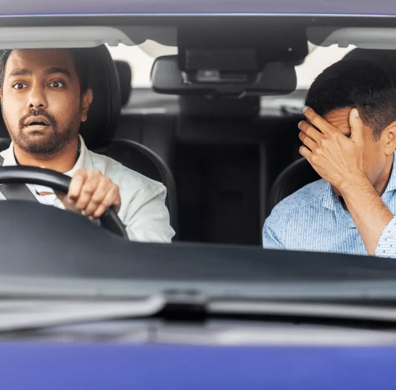 Worried driver sitting behind the wheel while a passenger covers his face after a driving incident, illustrating an insurance claim situation.