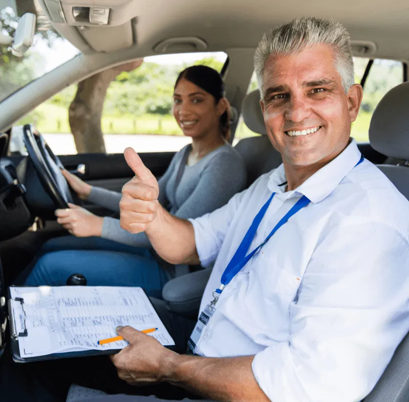 Driving instructor in passenger seat smiles and gives thumbs up while student driver holds the steering wheel during a lesson.