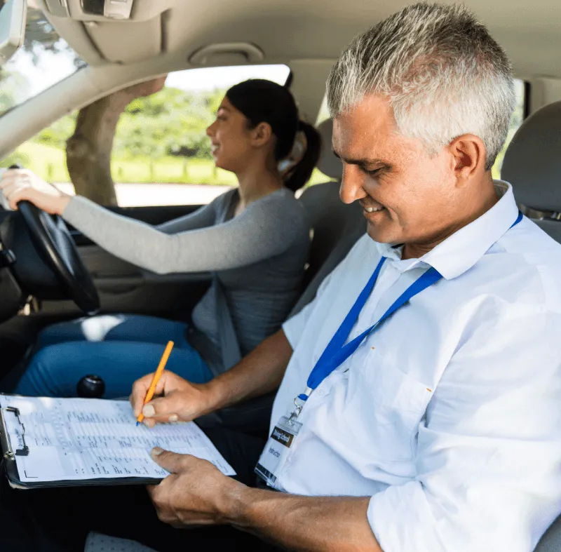 Driving instructor smiling while marking a checklist as a student practices driving a car.