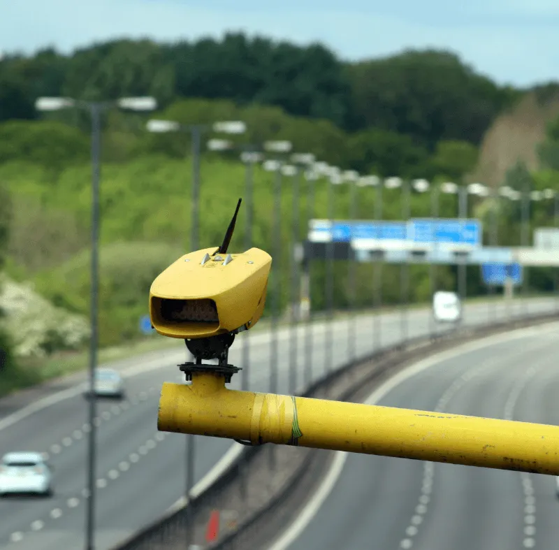 Yellow speed camera mounted on a yellow pole overlooking a UK motorway with street lights and blue road signs in the background.
