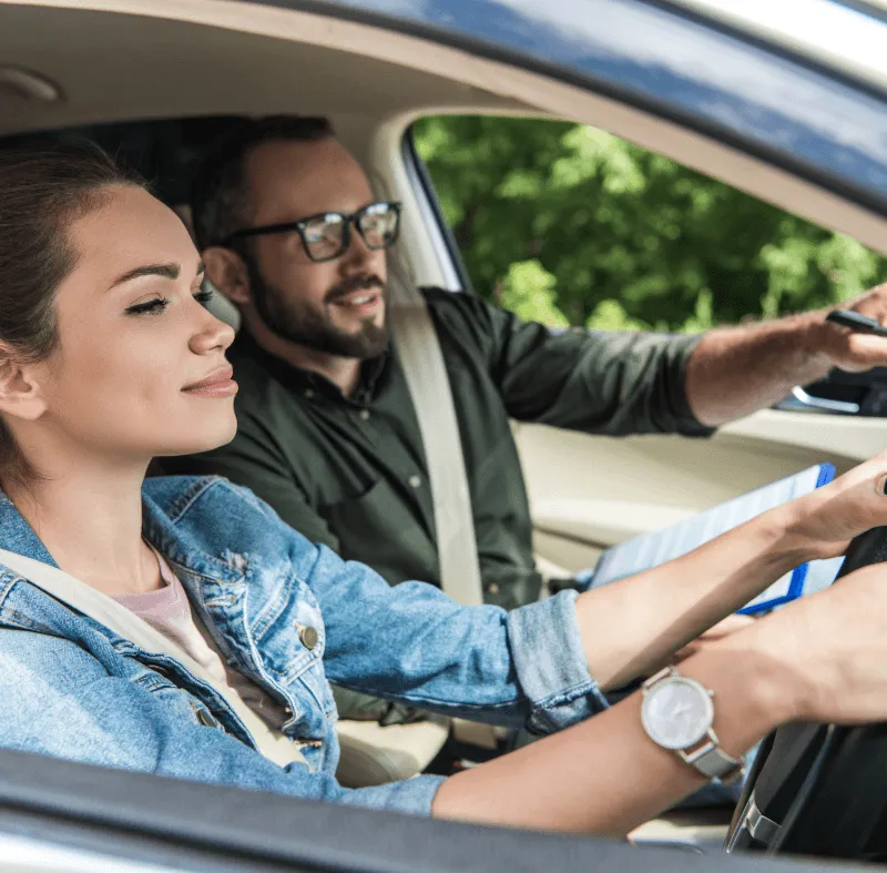 Young woman at the steering wheel during a driving lesson while a male instructor points and gives directions inside a car.