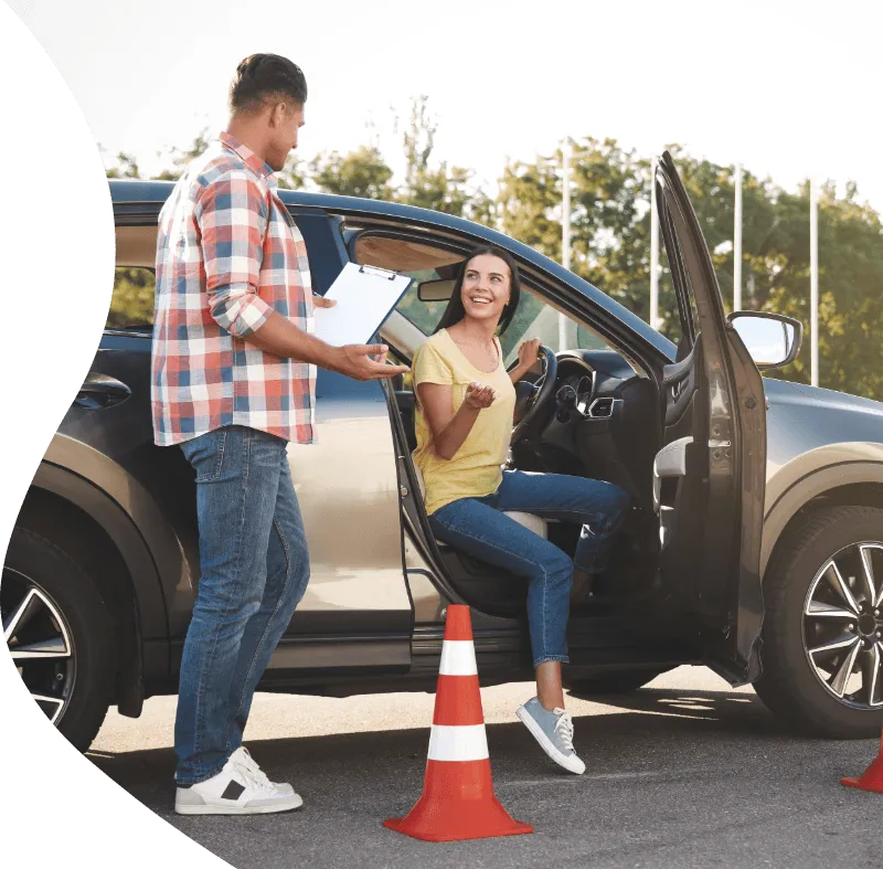 Driving instructor with clipboard talks to smiling woman in car during a lesson, surrounded by traffic cones in a parking lot.