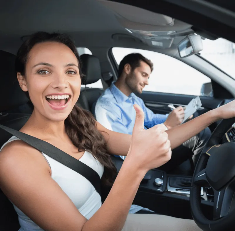 Young woman smiling and giving thumbs up while driving with a male instructor taking notes during a driving lesson.