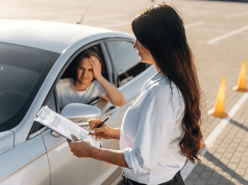 A driving examiner with a clipboard reviews notes beside a car, while an anxious student driver sits inside near orange cones.