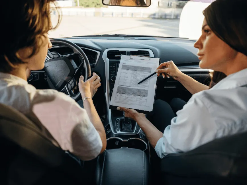 A driving instructor holding a clipboard reviews a form with a student driver seated behind the steering wheel inside a car.