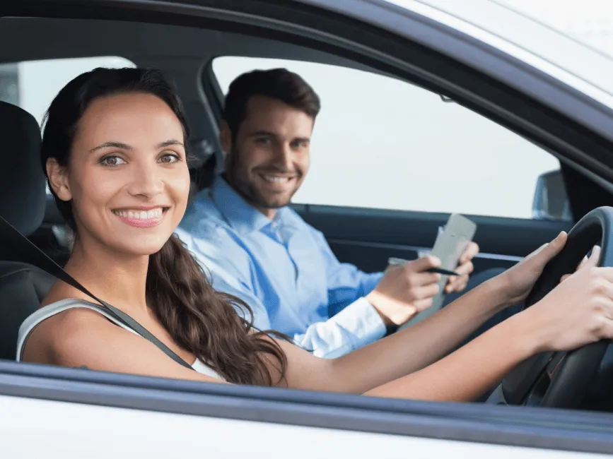 Smiling woman driving a car with a man in the passenger seat holding a smartphone, both looking at the camera.