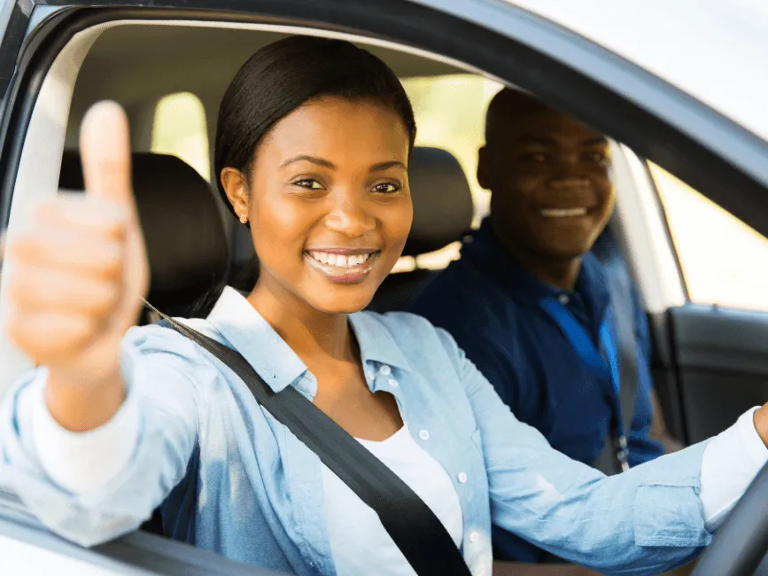 Smiling young woman in driver's seat giving thumbs up with seatbelt on, man in passenger seat, both happy inside a car.