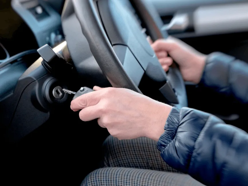 Person in a blue jacket inserting a car key into the ignition while holding the steering wheel inside a vehicle.