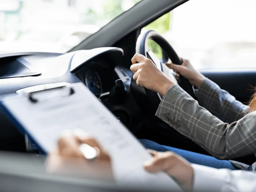 A student driver grips the steering wheel while an examiner holds a clipboard during a driving test evaluation.
