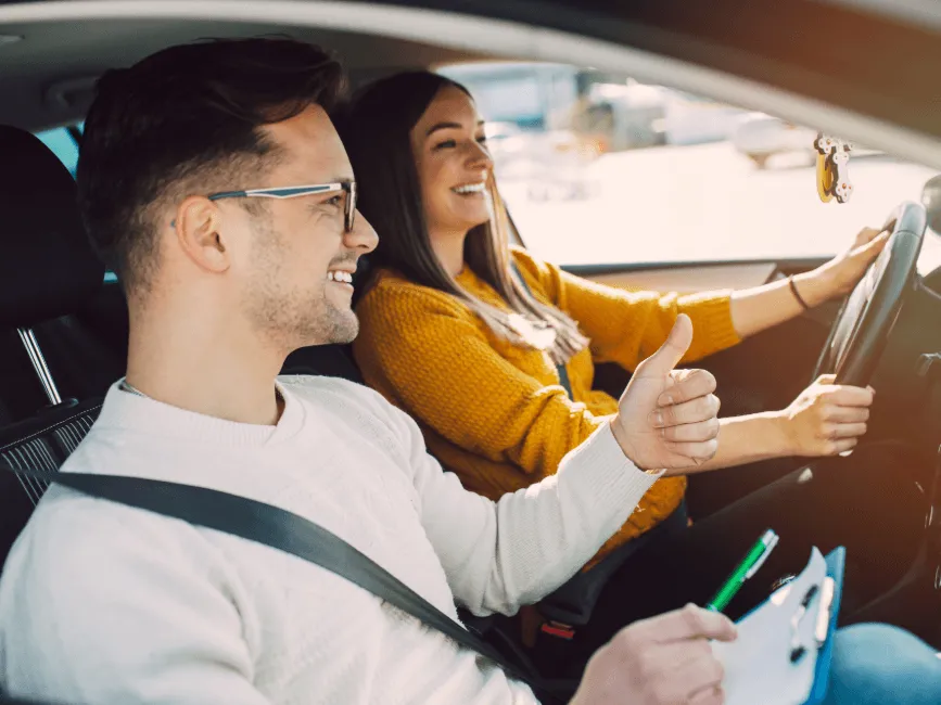Driving instructor giving thumbs up to smiling female student behind the wheel during a driving lesson in a car.