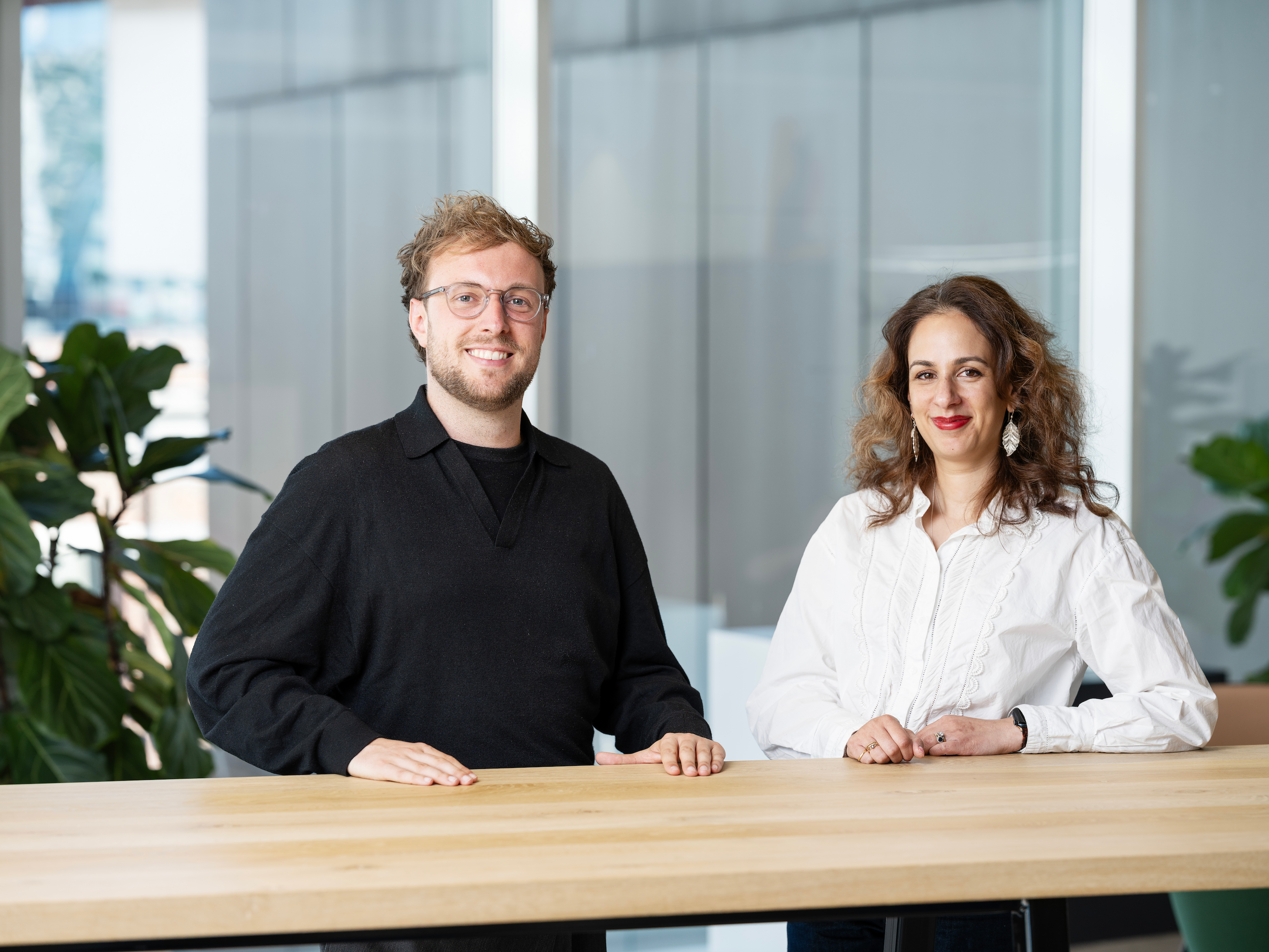 Two people smiling, standing beside a wooden table in a well-lit office with plants in the background.