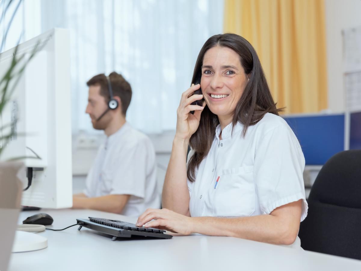 Mitarbeiterin am Empfang telefoniert lächelnd, Kollege mit Headset arbeitet im Hintergrund