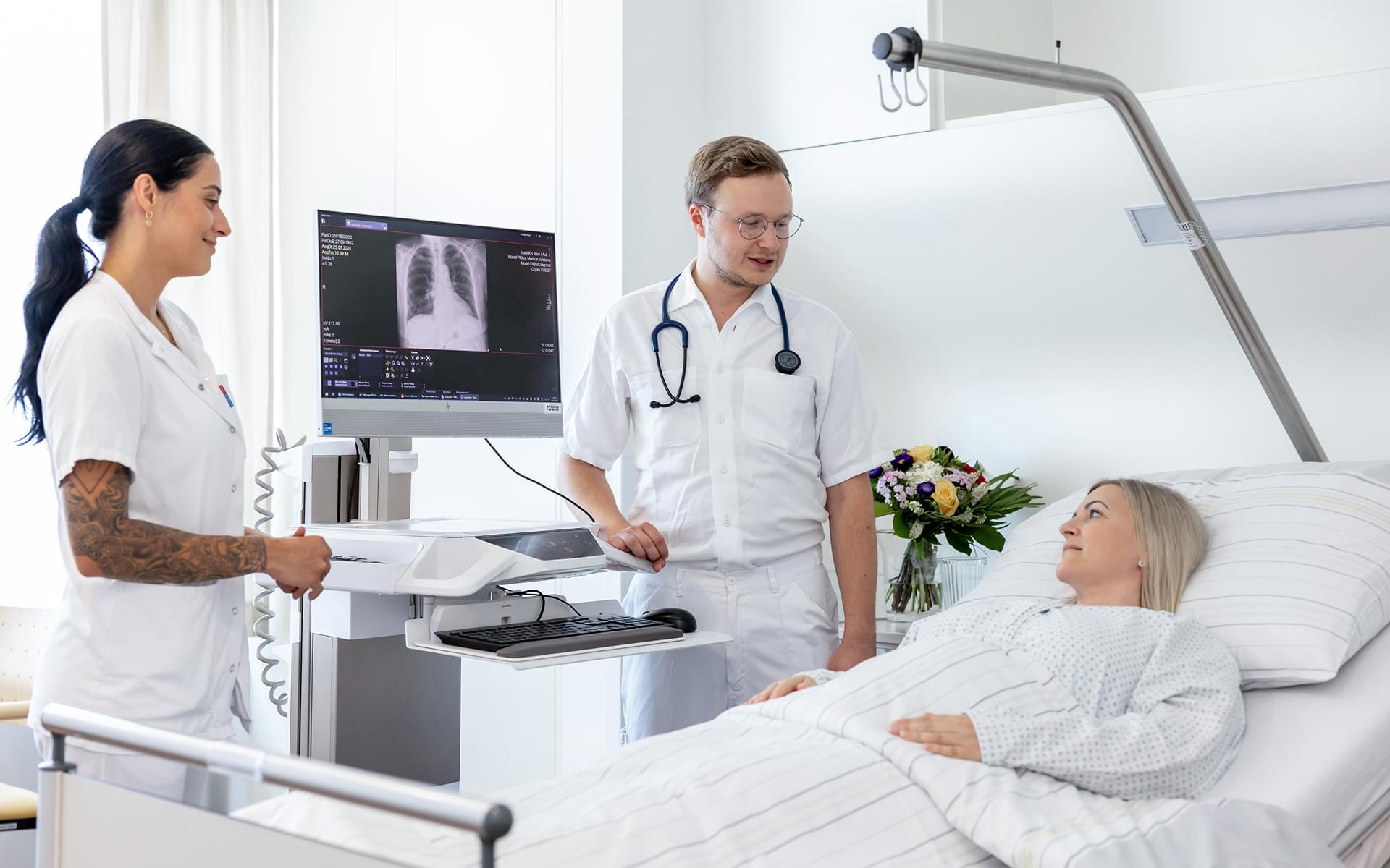 Ärztin und Arzt besprechen Röntgenbild am digitalen Bildschirm neben einer Patientin im Krankenbett