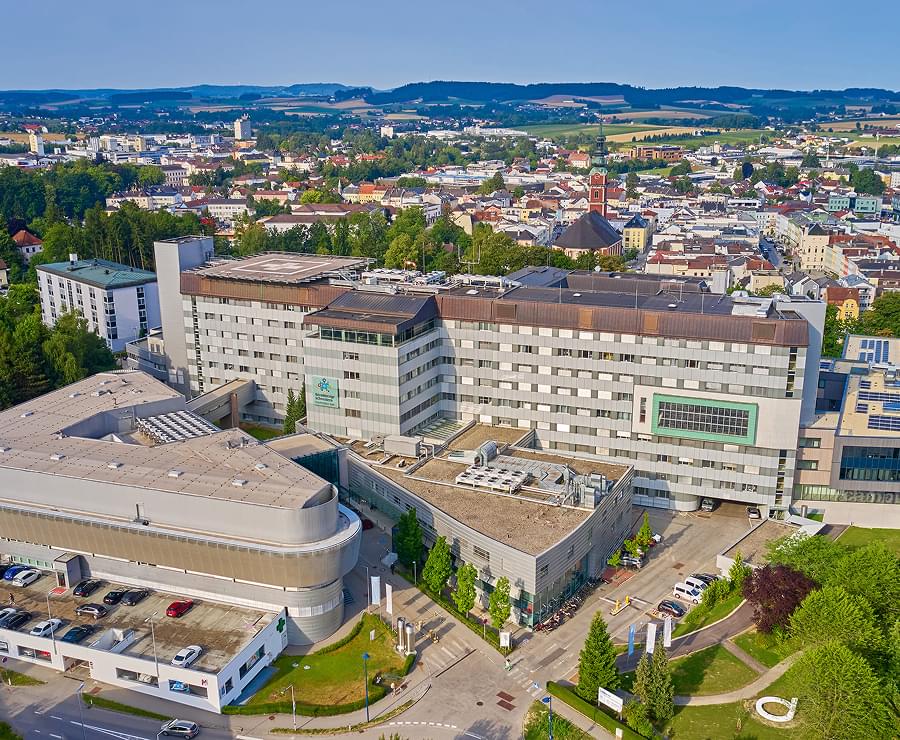 Luftaufnahme des modernen Krankenhauskomplexes BHS Ried mit Stadtpanorama im Hintergrund