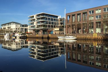 Ein Segelschiff liegt an einem Hafenbecken, das Wasser spiegelglatt, moderne Gebäude spiegeln sich darin.