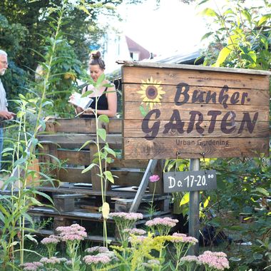 Eingangsschild des Bunkergartens, dahinter ein Einblick in den Garten.