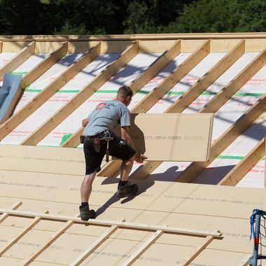Ein Arbeiter steht auf der offenen Holzkonstruktion eines Daches und verlegt Platten zur Dämmung.