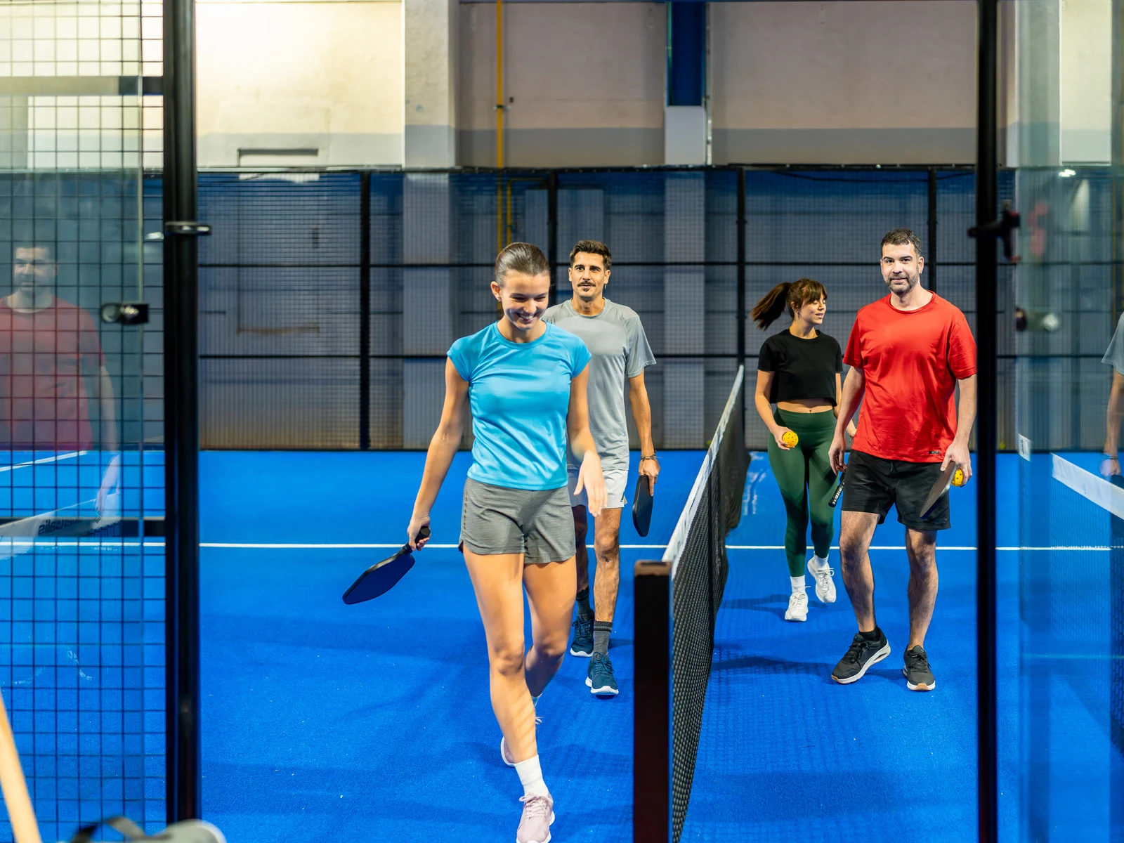 Padelhalle mit blauem Padel-Kunstrasen, Glaswänden und Spielern am Netz