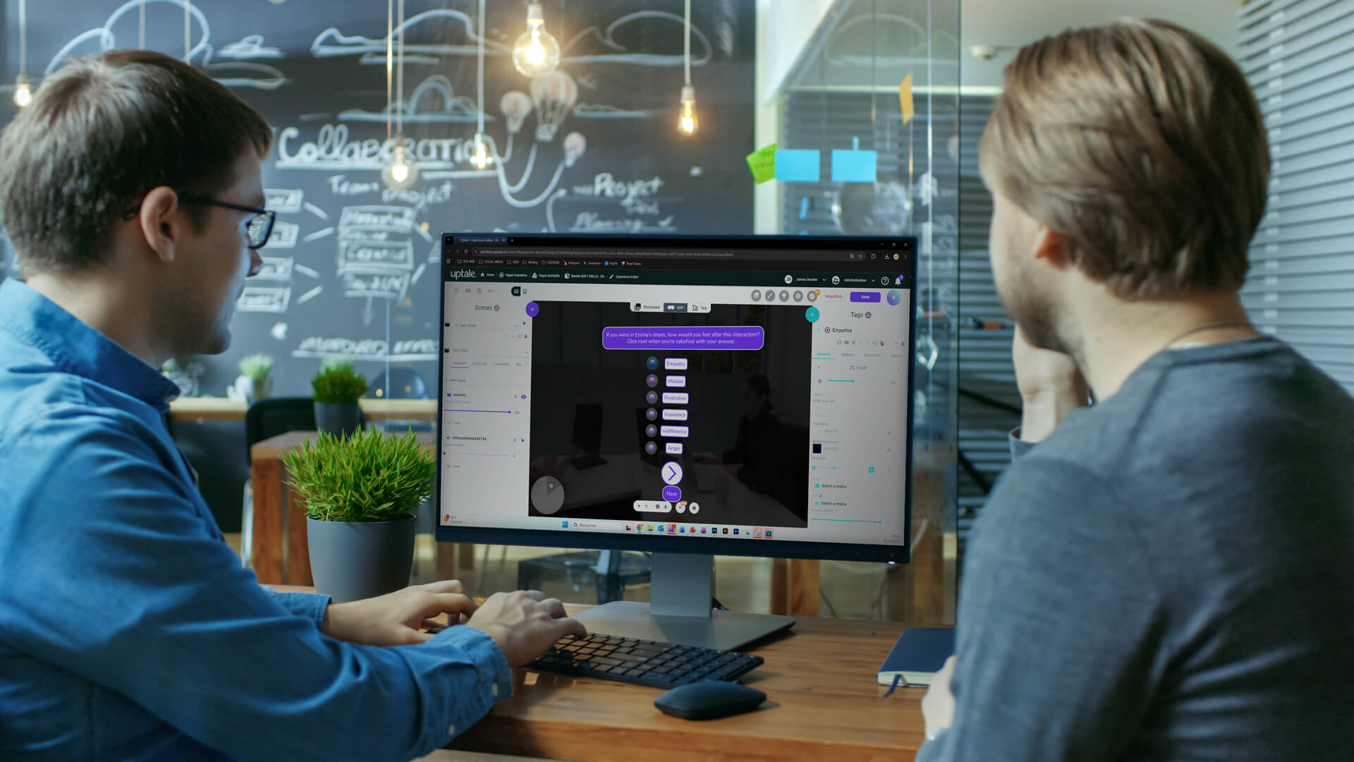 Two men sitting at a desk, collaborating on a project with a computer screen displaying a flowchart. A chalkboard wall is in the background.