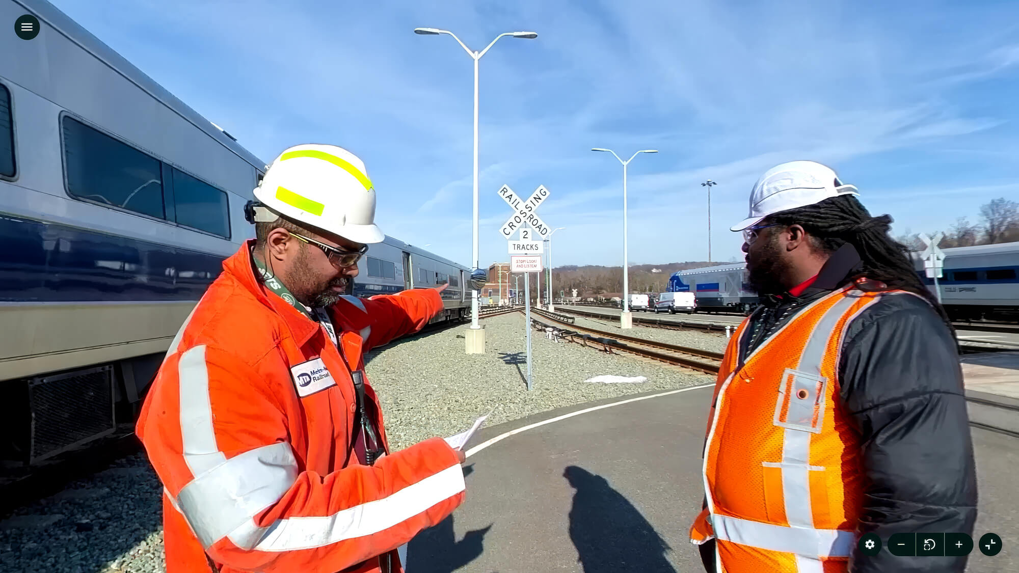 Two workers in orange safety vests and helmets discuss near a railway crossing with trains in the background on a clear day.