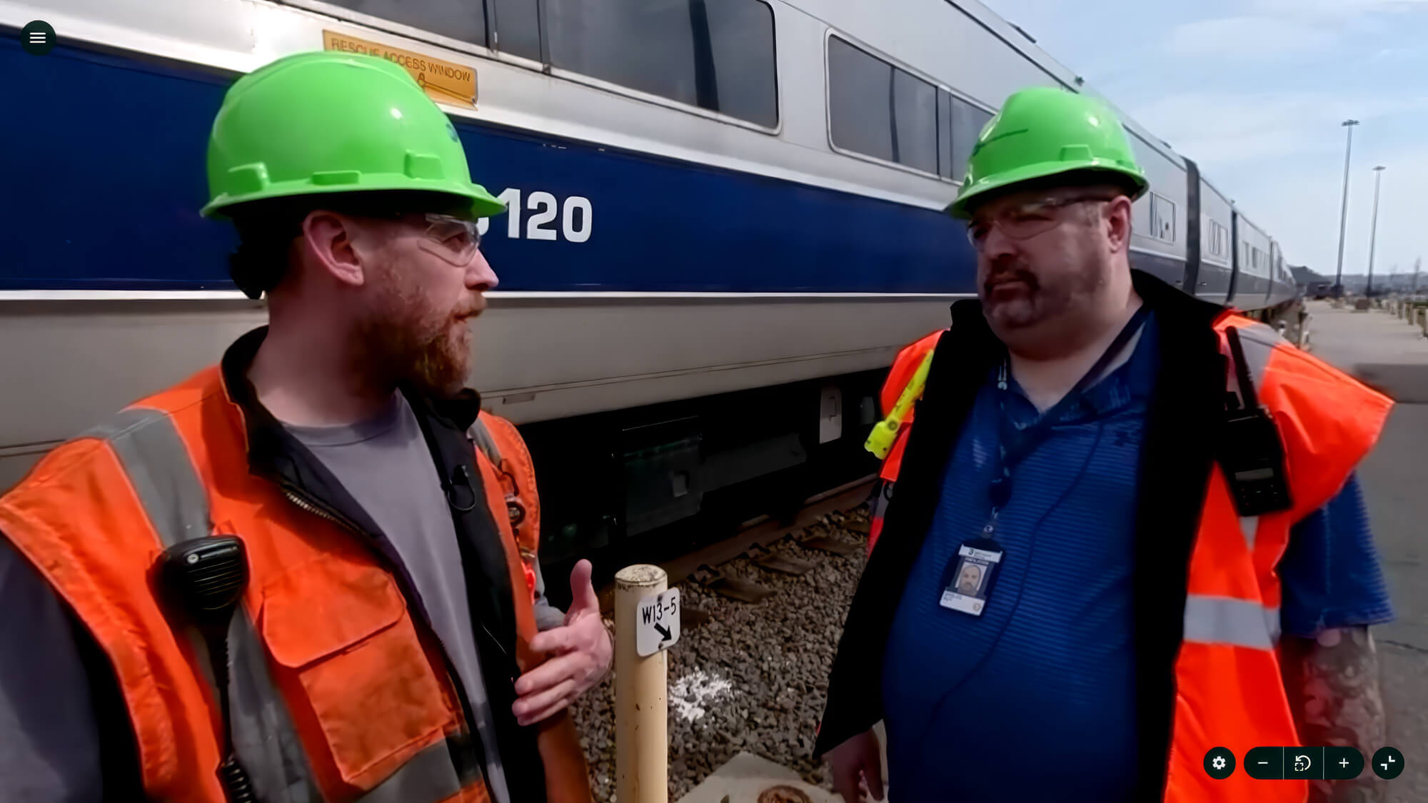 Worker in safety gear inspects between train cars on railway tracks, wearing a green helmet and orange vest.
