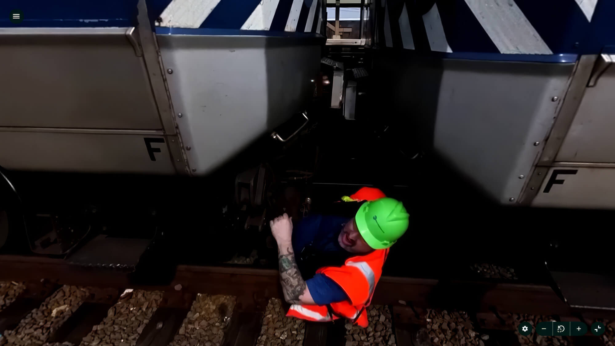 Worker in safety gear inspects between train cars on railway tracks, wearing a green helmet and orange vest.