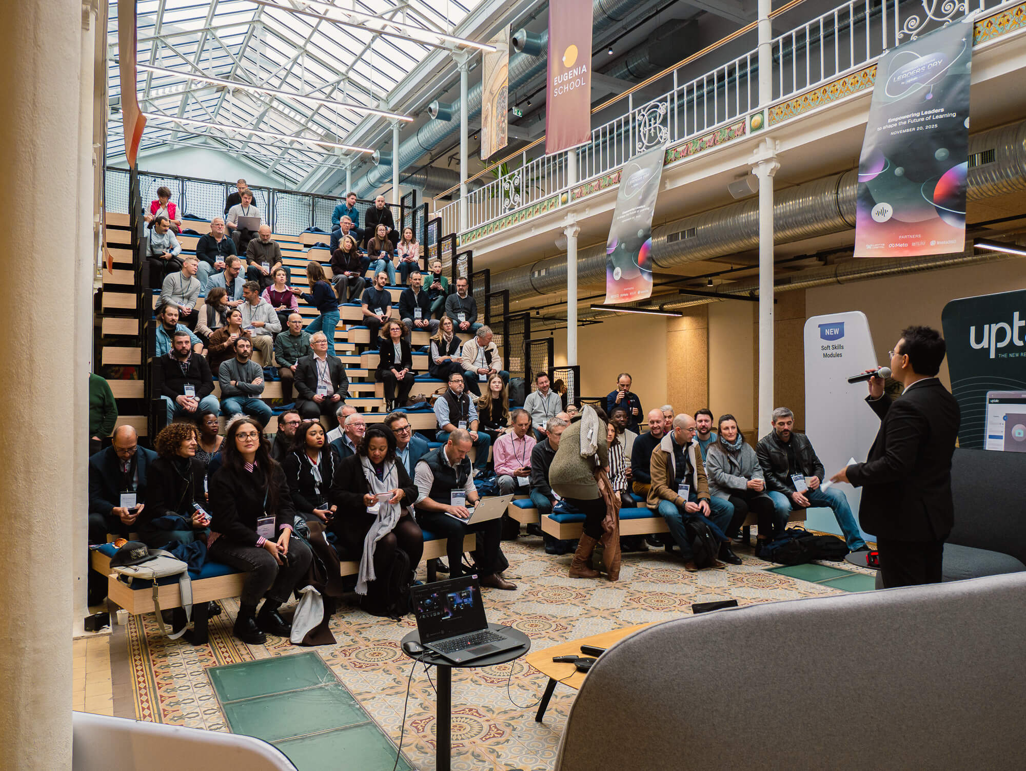 A person speaks to a diverse audience seated in a modern, open space with a high glass ceiling and banners displaying event information.