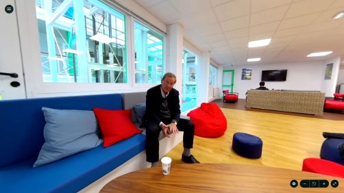 A person sits on a blue couch in a bright lounge area with red cushions, bean bags, and wooden flooring. A TV is mounted on the wall.