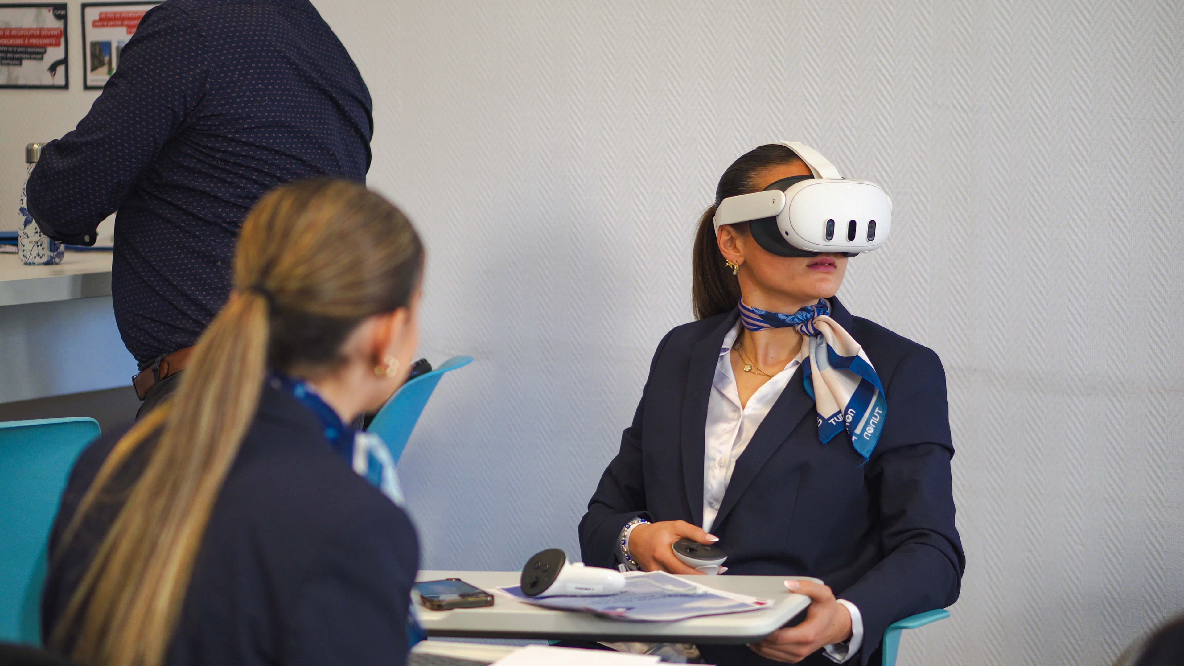 A woman in formal attire, wearing a VR headset, sits with a controller in her hand. Another woman with a ponytail sits nearby.