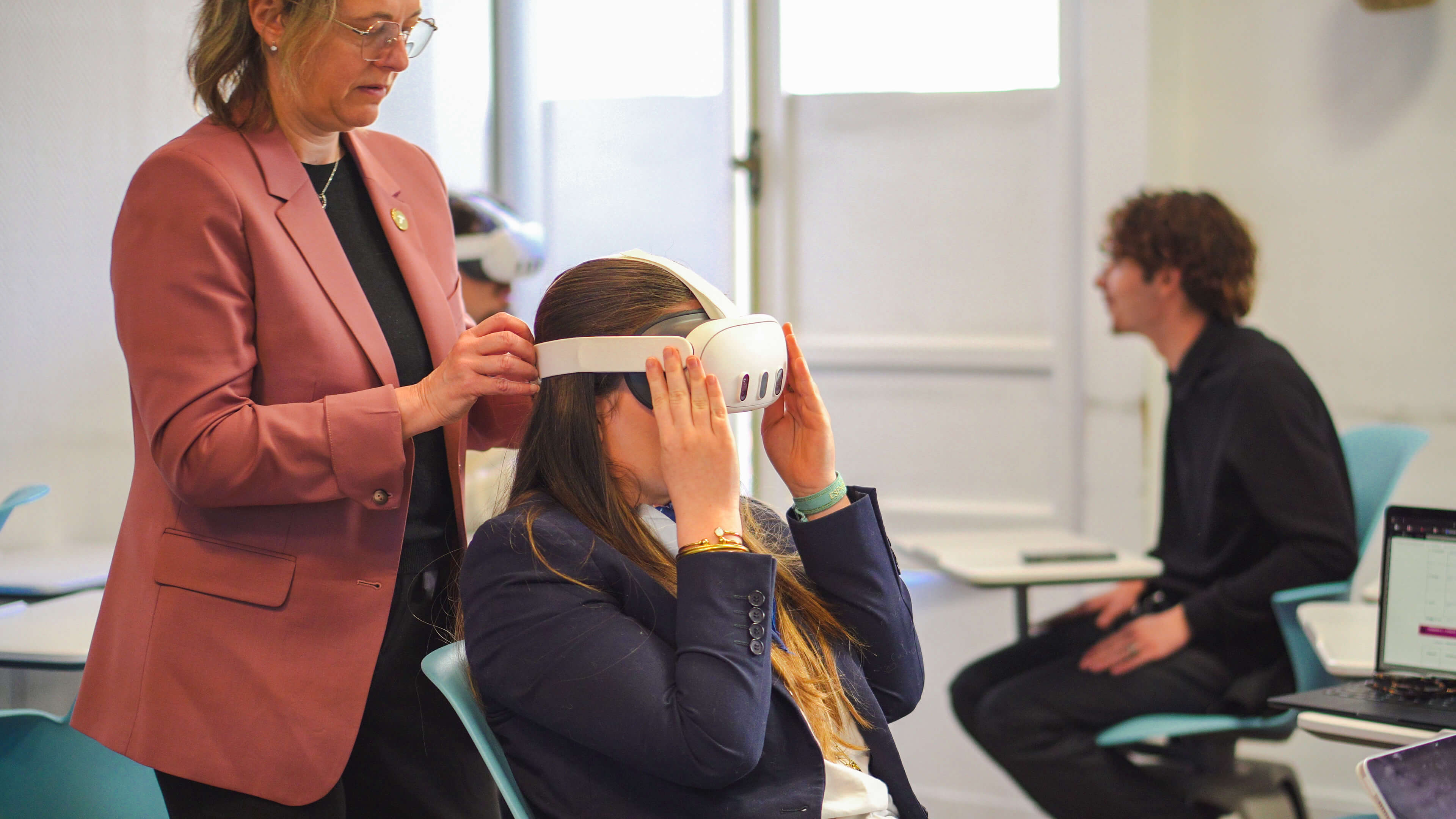 A woman adjusts a VR headset on a seated person's head in a classroom. Another person sits in the background by a laptop.