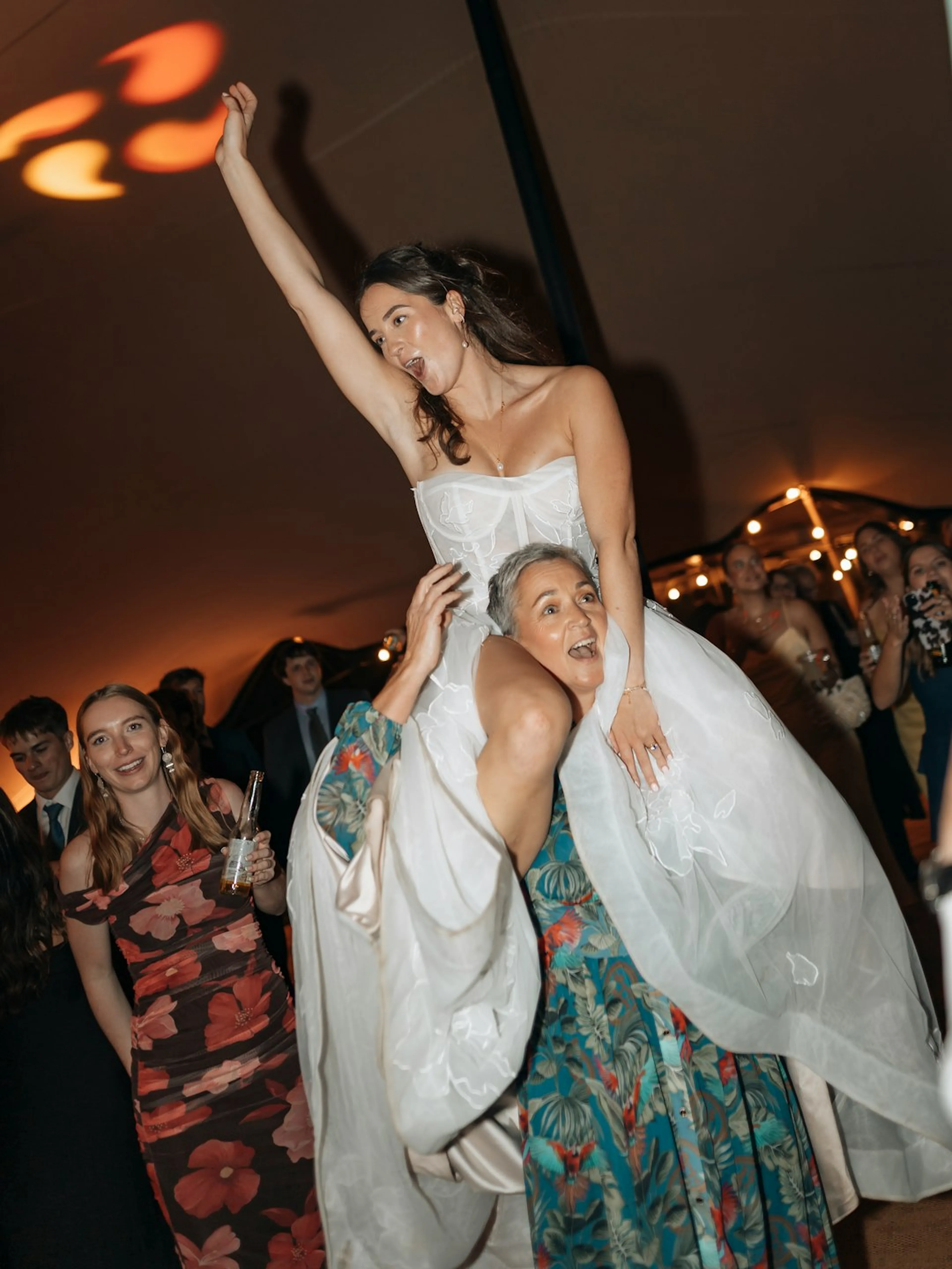 A bride lifted mid-cheer under string lights at a wedding reception