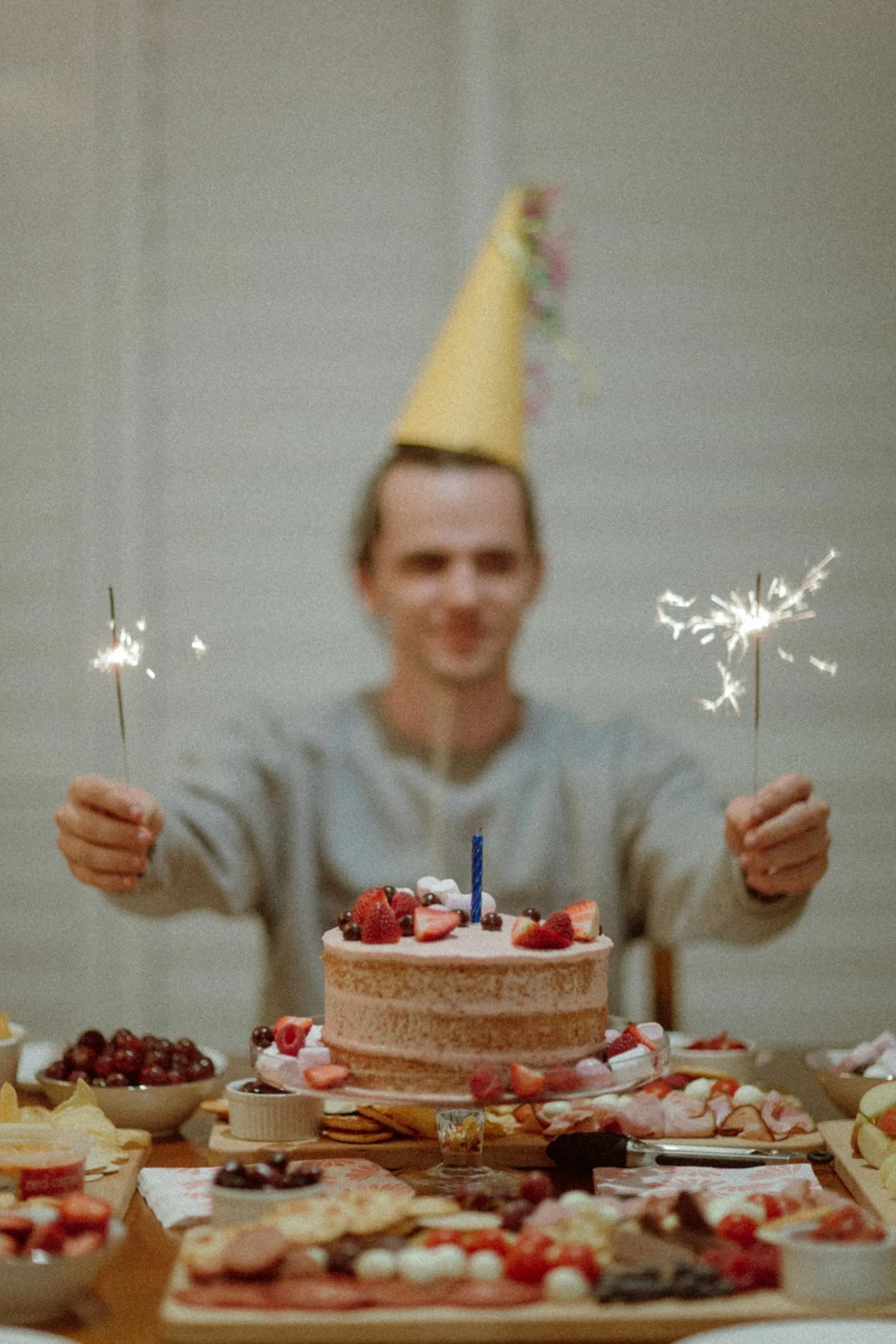 A person in a party hat holding sparklers behind a birthday cake