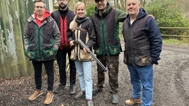 Five people in outdoor gear pose together, one holding a shotgun, next to a corrugated metal wall in a woodland setting.