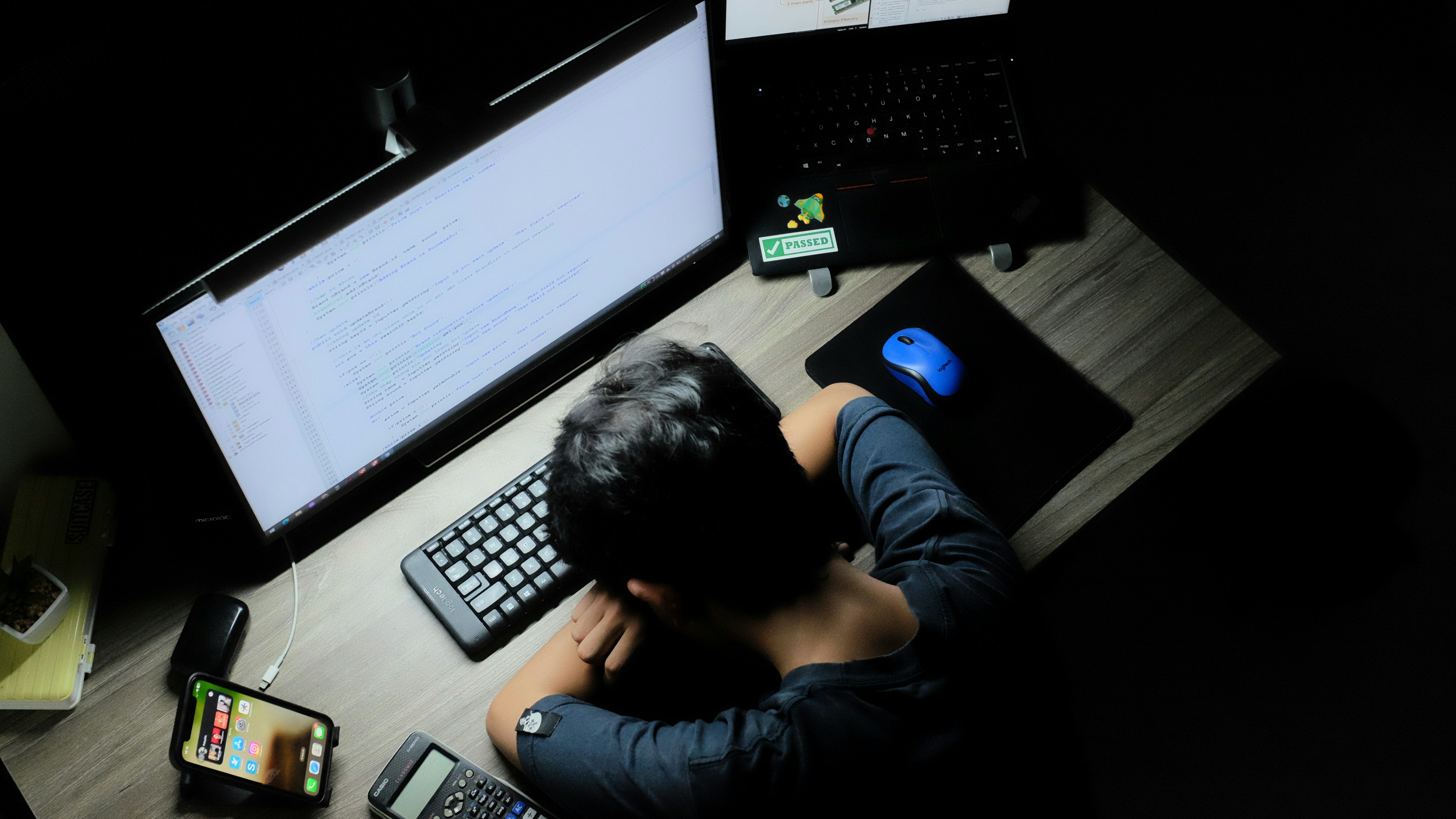 A person wearily rests their head on their arms at a desk with a computer.