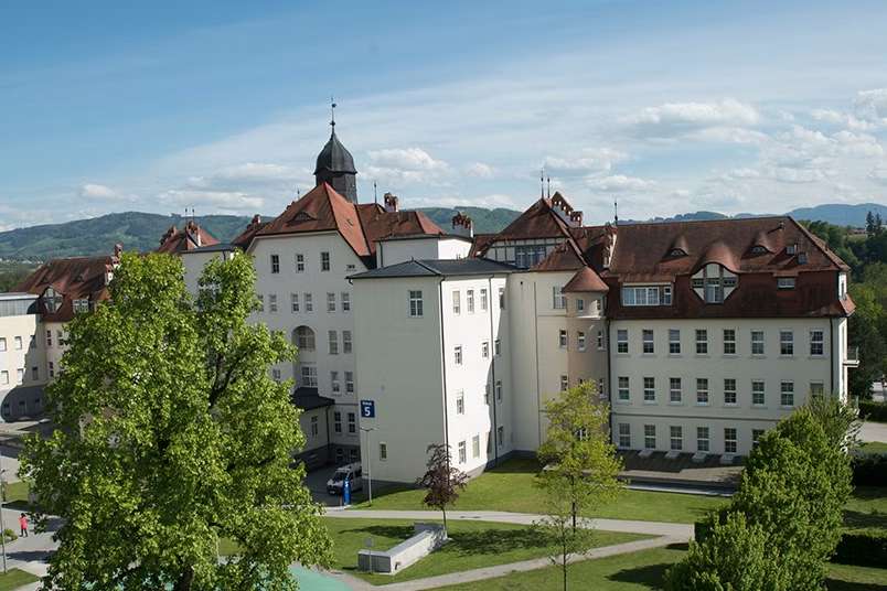 Gebäude mit vielen Fenstern und prunkvoller Dachoptik mit davor liegender Parkfläche.