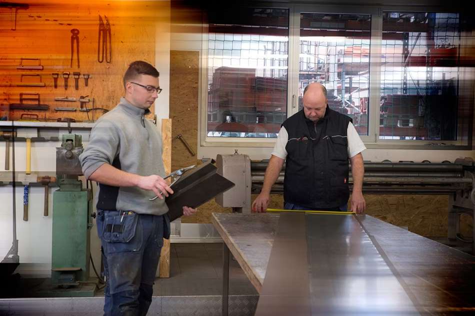 Zwei Männer in Arbeitskleidung beim Vermessen und Anschneiden von Baumaterialien in einer Werkstatt.