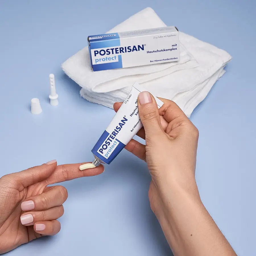 Hände tragen Posterisan protect Salbe aus der Tube auf einen Finger auf. Im Hintergrund befindet sich der Applikator und die Verpackung auf gefalteten Handtüchern.