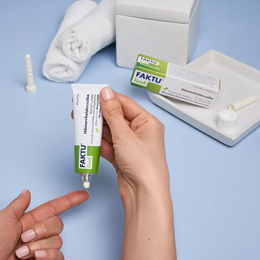 FAKTU lind Hämorrhoidensalbe wird aus einer Tube auf einen Finger aufgetragen; im Hintergrund liegen die Produktverpackung, der Applikator und gerollte weiße Handtücher.